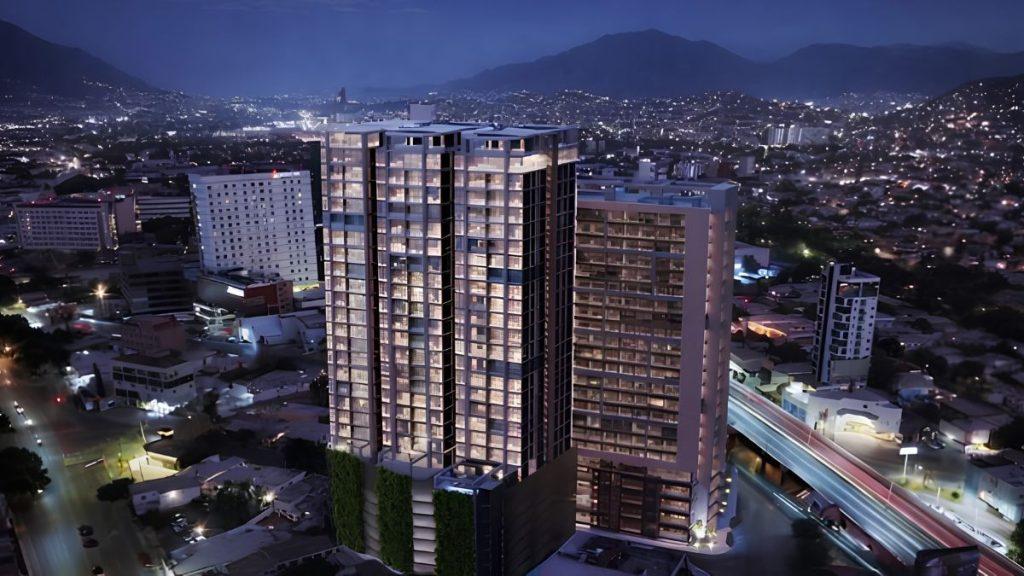Aerial night view of two modern glass residential towers illuminated in a city with glowing streets and distant mountains in the background.