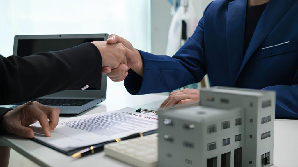 Two professionals shake hands over a signed contract on a desk, with a laptop and a miniature building model nearby.