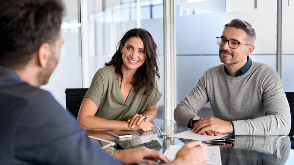 Three professionals sit at a glass conference table, smiling during a meeting.